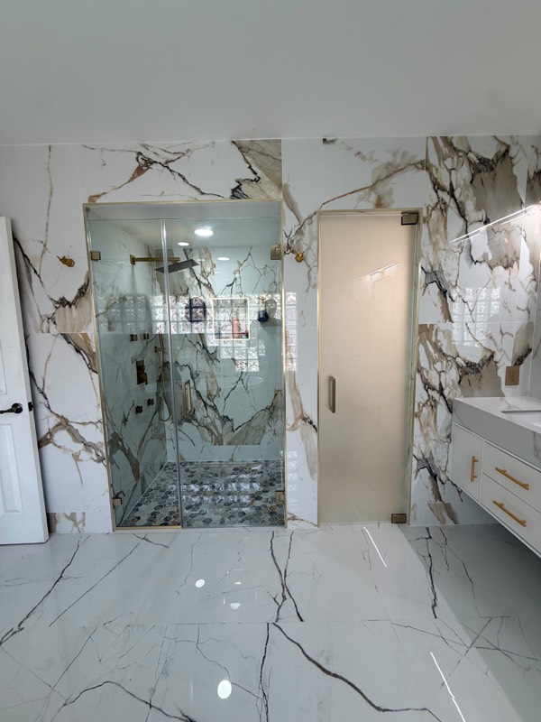 Frameless shower enclosure with gold trim next to a frosted glass door in a marble bathroom